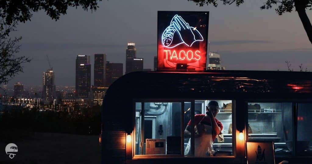 Taco food truck owner giving the peace sign from his food truck which is parked on the hill overlooking the los angeles skyline at night time.