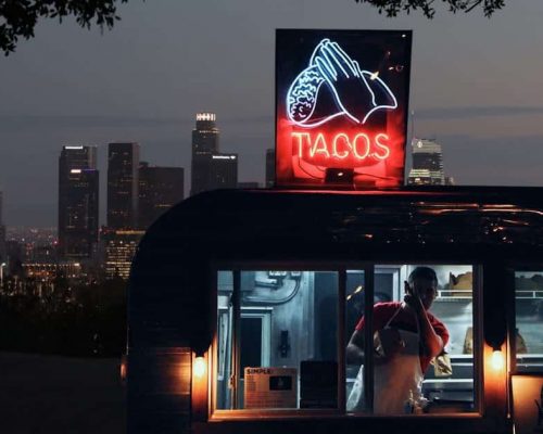Taco food truck owner giving the peace sign from inside his truck while parked on a hill overlooking los angeles skyline at night